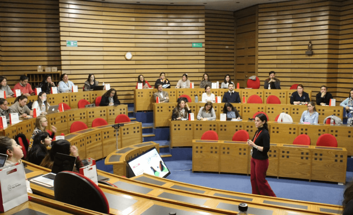 Una mujer de pie en una sala de conferencias con paredes de madera, hablando frente a un grupo de personas sentadas en filas de asientos elevados con respaldos rojos. Las personas están atentas, tomando notas y usando computadoras portátiles.
